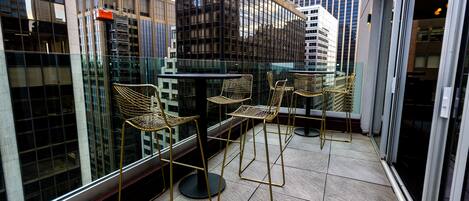 A balcony with four wicker chairs and a small table, overlooking a cityscape with tall buildings.