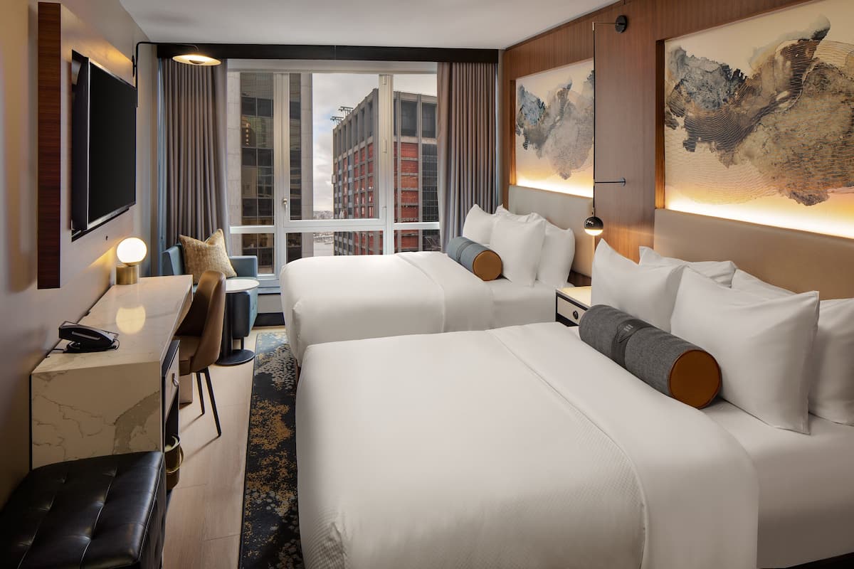 A hotel room with two beds, a desk, a chair, a TV, and a large window with a city view.
