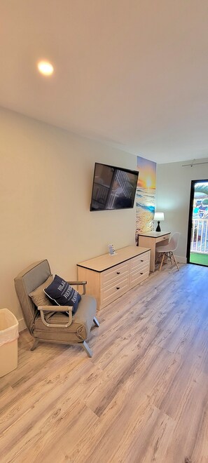 Een kamer met een houten vloer, een bureau met een lamp, een stoel, een aan de muur bevestigde televisie en een raam met uitzicht op een omheining.