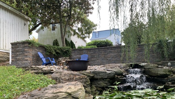 A backyard with a small waterfall, rocks, and a pond with lily pads. There are two blue chairs and a black pot near the waterfall. - Turnbridge Manor (Denton)