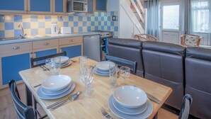 A compact kitchen with blue cabinets, a microwave, and a stove. There is a dining area with a wooden table set for four, including plates, glasses, and cutlery. - Trewent Park - 2 Bed - Freshwater East (Pembroke)