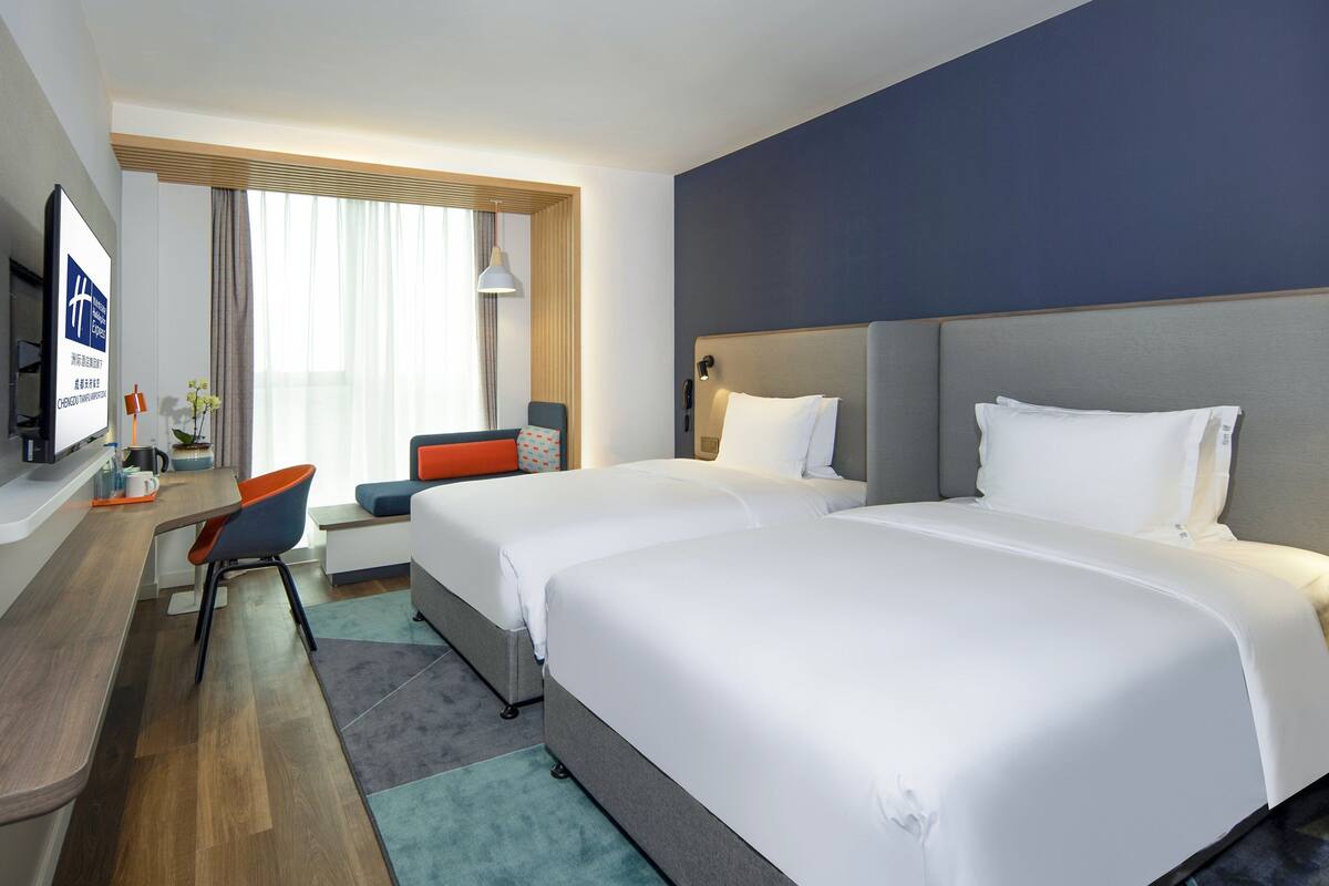 A hotel room with two beds, a desk with a chair, a TV, and a window with curtains.