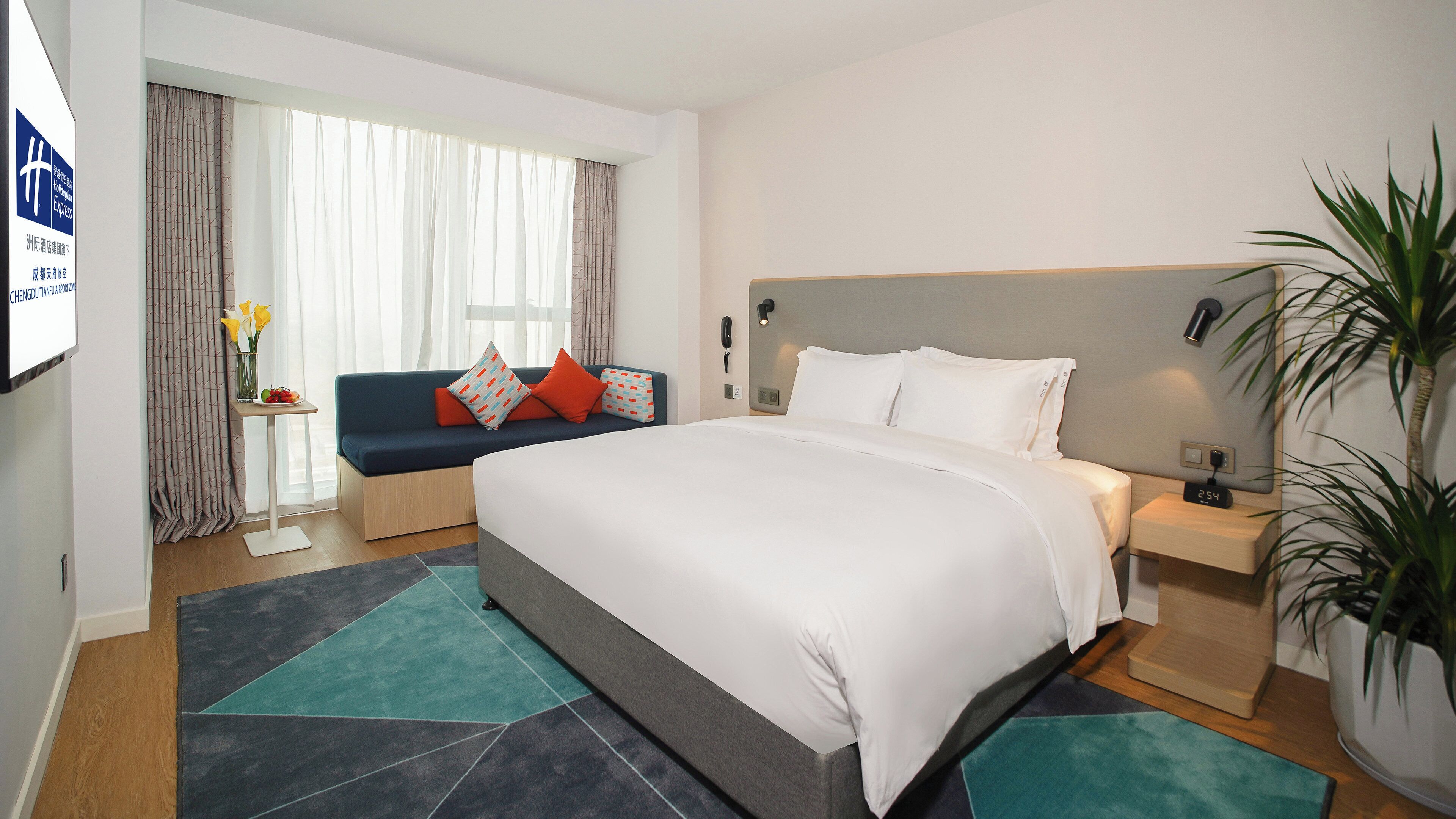 A hotel room with a large bed, a bedside table with a lamp, a sofa with cushions, a small table with a vase of flowers, and a large window with curtains.