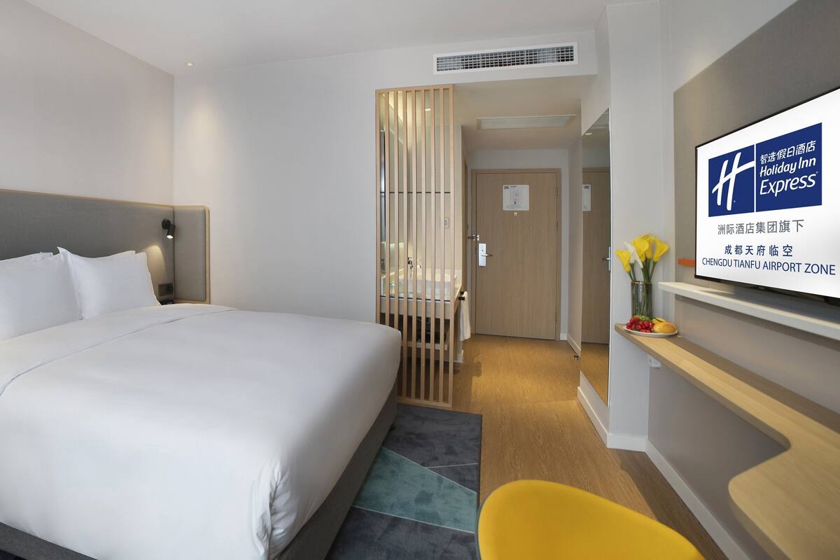 A hotel room with a large bed, a nightstand, a desk with a chair, a TV on the wall, and a decorative vase with flowers.