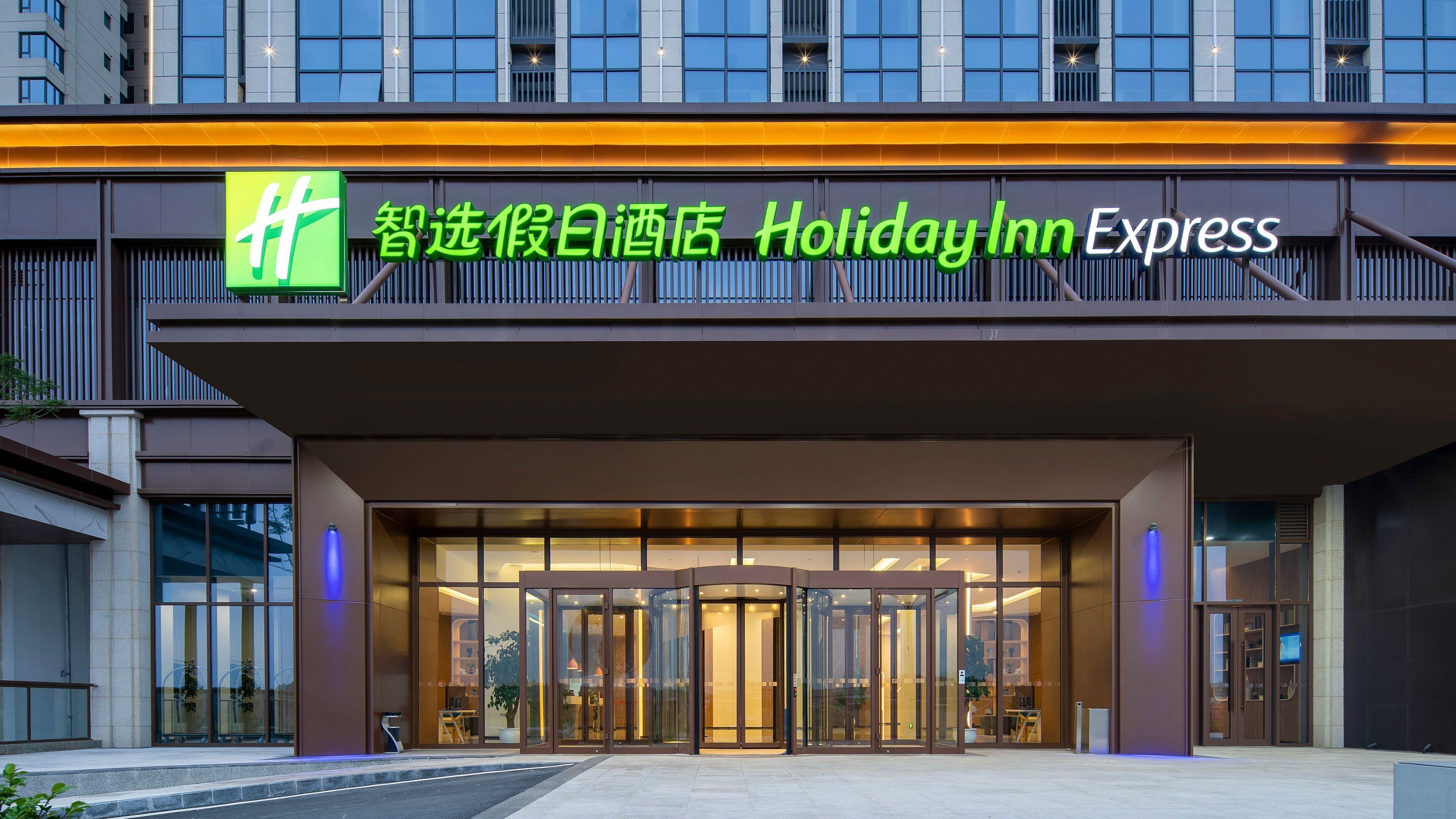 Entrada de un hotel Holiday Inn Express con diseño moderno, destacando un logotipo verde prominente, fachada de vidrio y un conjunto de puertas giratorias.