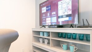 A living room with a flat-screen TV displaying a streaming service interface, a white shelving unit with various mugs, and a modern chair.