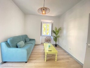A room with a light blue sofa, a yellow table, a hanging light fixture, a window with blinds, and a potted plant. - Setúbal Downtown Todi II (Setúbal)