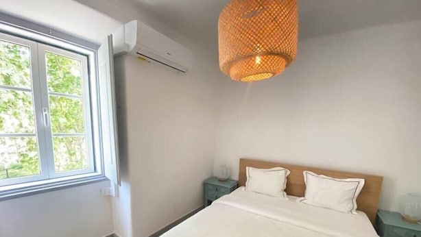 A bedroom with a bed, two nightstands, an air conditioning unit, and a window with a view of greenery. - Setúbal Downtown Todi II (Setúbal)