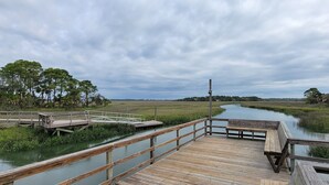 A wooden boardwalk leading to a small dock over a calm water body, surrounded by grassy areas and trees. - First Floor Beach Club Villa with Marsh Views - Walk to the Beach! (Saint Helena Island)