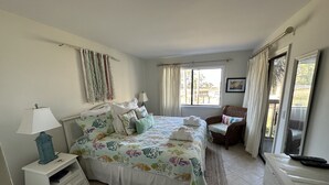 A bedroom with a bed, bedside table, chair, and a window with curtains. - First Floor Beach Club Villa with Marsh Views - Walk to the Beach! (Saint Helena Island)