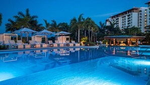 Ein Schwimmbecken mit Liegestühlen und Sonnenschirmen, umgeben von Palmen und einem Hotelgebäude.