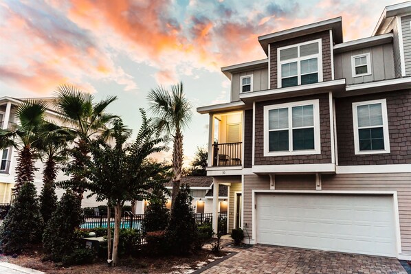 A multi-story residential building with a garage, a balcony, and a pool in the background.