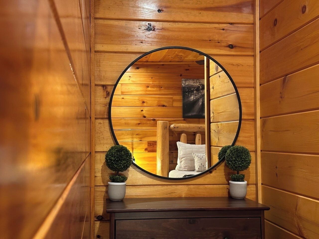 Ein Holzblockhaus-Innenraum mit einem runden Spiegel, der ein Bett und zwei Blumentöpfe reflektiert.