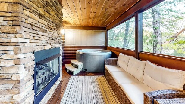 L’intérieur chaleureux d’un chalet avec une cheminée en pierre, un bain à remous et un canapé placé près d’une grande fenêtre donnant sur la forêt.