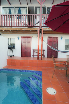Une piscine aux carreaux bleus, un parasol rouge et une terrasse en bois avec un hamac.
