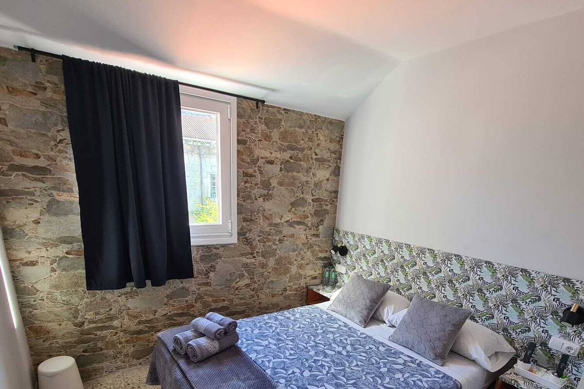 A bedroom with a stone wall, a window with curtains, a bed with a patterned bedspread, and a nightstand with a lamp.