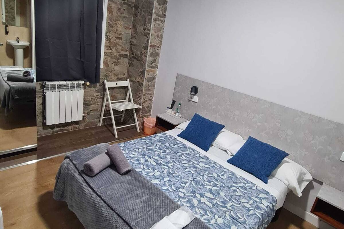 A modern bedroom with a large bed, a small desk, a chair, and a bathroom visible through a glass door.