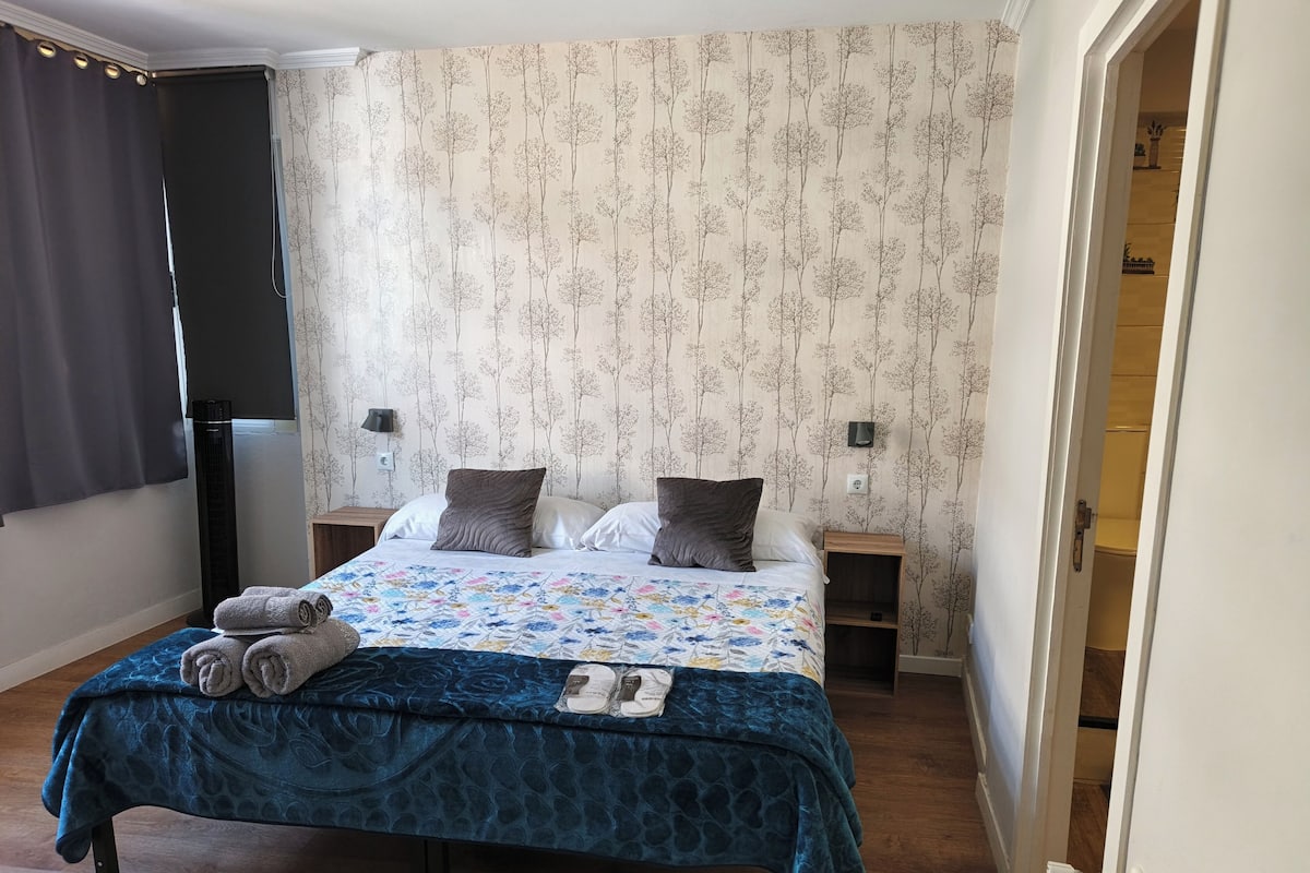 A bedroom with a bed, two bedside tables, a radiator, a window with curtains, and a door to another room.