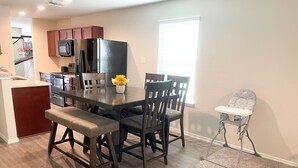 A modern kitchen with dark wooden cabinets, a stainless steel refrigerator, and a microwave. There is a dining table with chairs and a small side table with a chair. A vase with flowers is on the table.