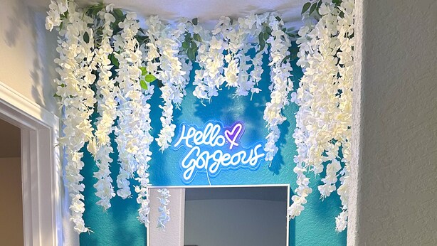 A teal wall with a mirror and a neon sign that reads "Hello Gorgeous."