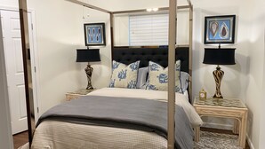 Un lit à baldaquin avec une couvre-lit grise, deux tables de chevet assorties avec des lampes, une fenêtre avec des stores et deux œuvres d’art encadrées accrochées au mur.