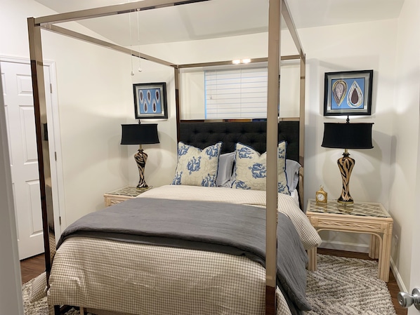 A four-poster bed with a grey bedspread, two matching nightstands with lamps, a window with blinds, and two framed artworks on the wall.