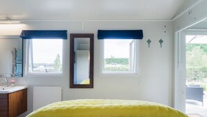 A bedroom with a bed, a mirror, two windows with blue curtains, a sink, and a view of greenery outside.