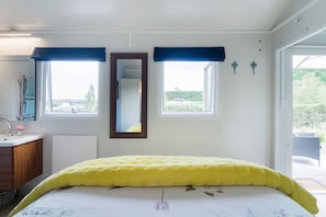 A bedroom with a bed, a mirror, two windows with blue curtains, a sink, and a view of greenery outside.