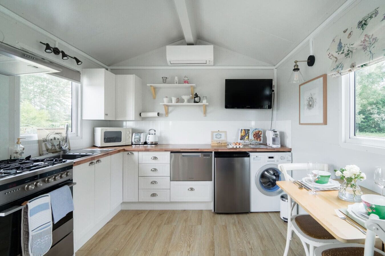 A modern kitchen with white cabinetry, a gas stove, a microwave, a dishwasher, and a washing machine. There is a wooden countertop, a dining table with chairs, and a window providing natural light.