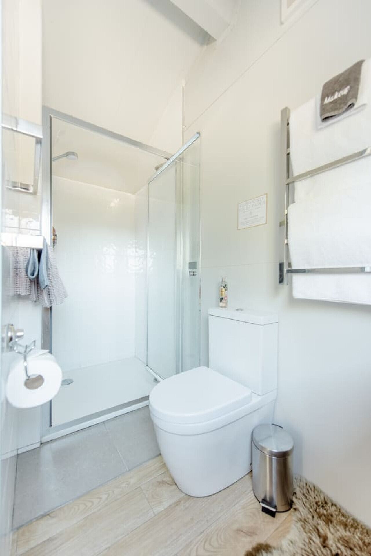 A modern bathroom with a glass shower enclosure, a toilet, a trash bin, and a towel rack.
