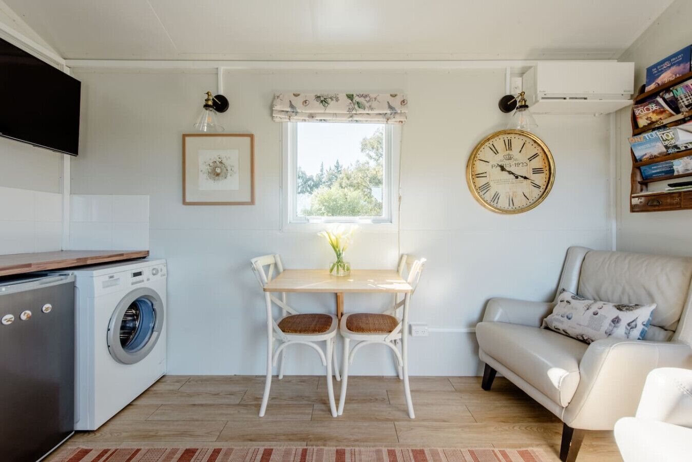 A compact living space with a washing machine, a dining table with chairs, a sofa, a clock, and a window with a view of greenery.