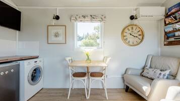 A compact living space with a washing machine, a dining table with chairs, a sofa, a clock, and a window with a view of greenery.