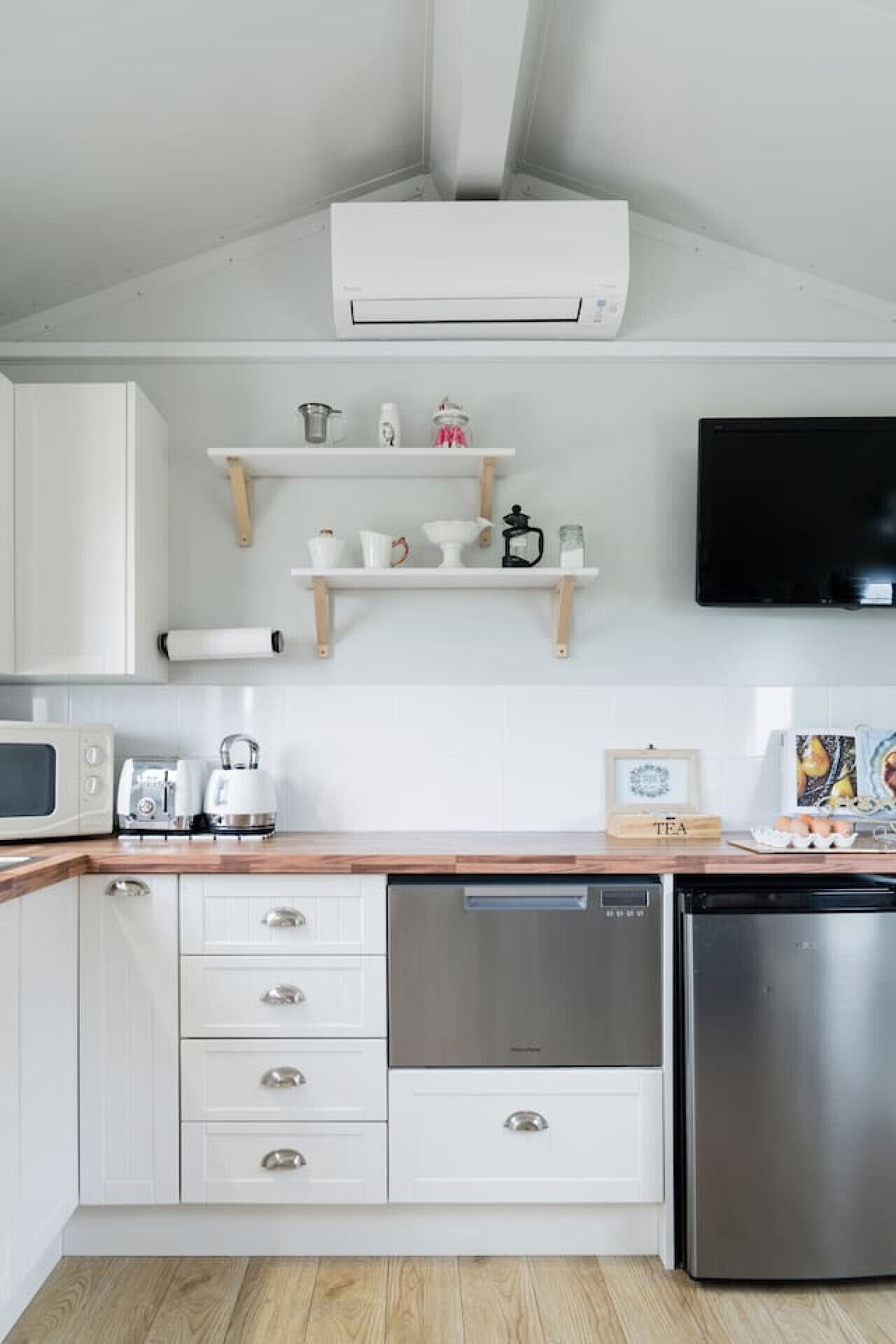 A modern kitchen with white cabinetry, a stainless steel dishwasher, a microwave, and a coffee maker. There are wooden shelves above the counter holding various kitchen items.