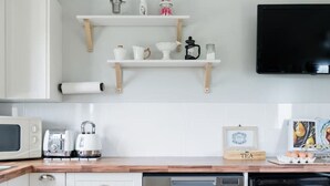 A modern kitchen with white cabinetry, a stainless steel dishwasher, a microwave, and a coffee maker. There are wooden shelves above the counter holding various kitchen items.