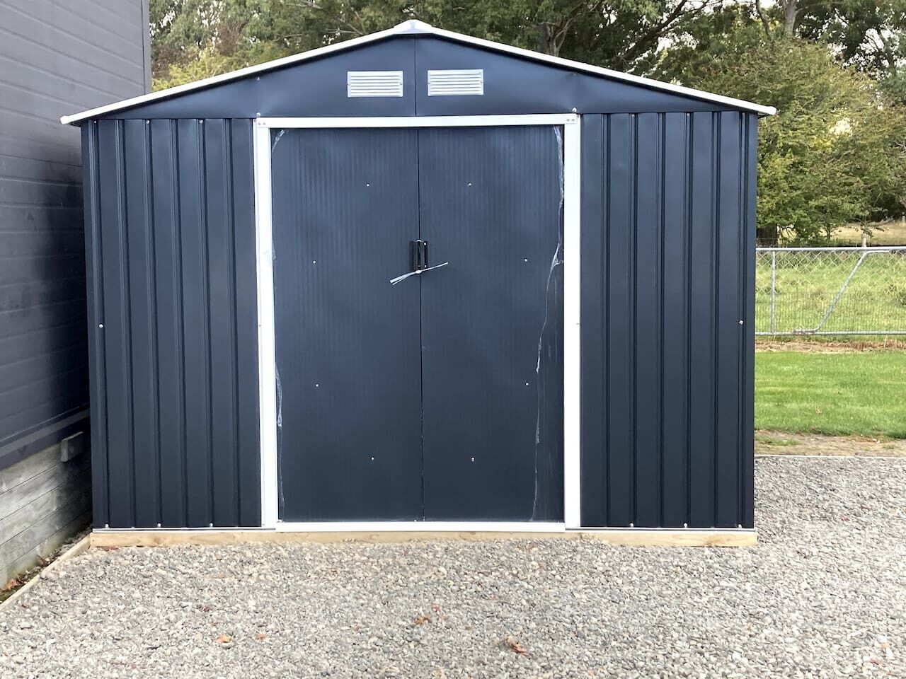 Eine dunkel gefärbte Gartenhütte mit weißer Tür und Lüftungsöffnungen am Dach.