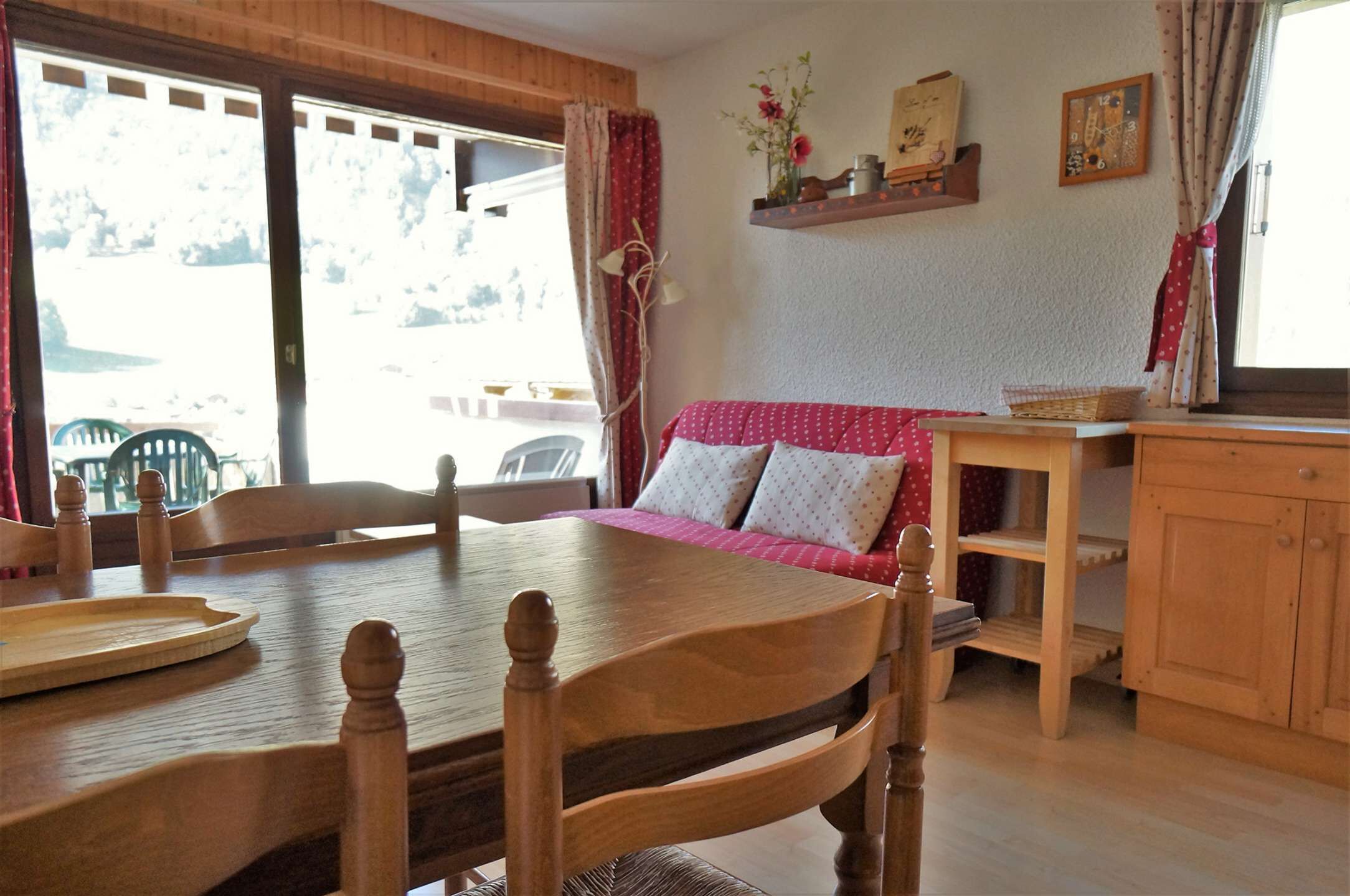 A wooden dining table with chairs, a red and white patterned sofa, and a wooden cabinet.