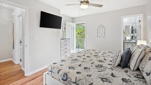 Un dormitorio con cama, ventilador de techo, televisor, cómoda y puerta que da a un balcón.