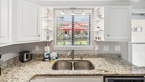 Una cocina con armarios blancos, encimera de granito, pileta doble y ventana con persianas.