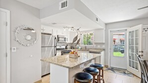 Una cocina moderna con armarios blancos, una isla central con taburetes, un reloj decorativo en la pared y vistas al patio trasero a través de una puerta de cristal.