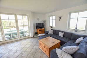 A spacious living room with a large grey sofa, a wooden coffee table, and a television on a wooden stand. There are large windows providing natural light and a view of the outside.