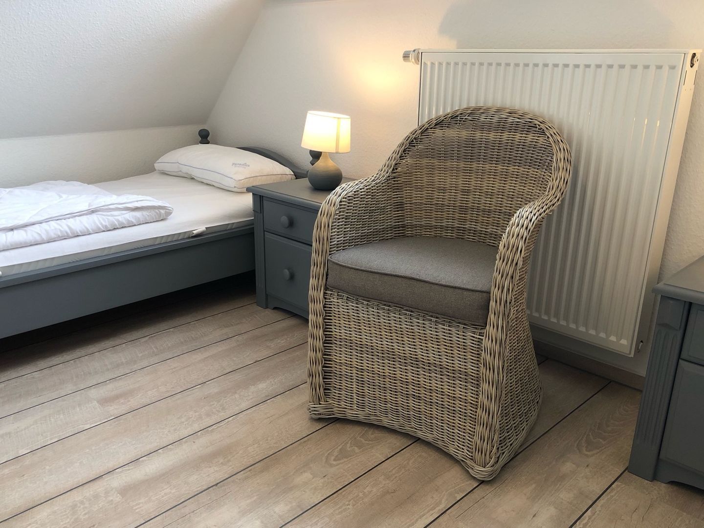 A bedroom with a bed, a nightstand, a lamp, a wicker chair, and a radiator.