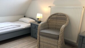 Une chambre à coucher avec un lit, une table de chevet, une lampe, un fauteuil en osier et un radiateur.