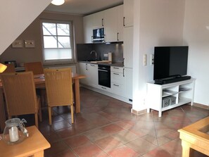 A compact kitchen with white cabinets, a microwave, oven, and a TV on a stand. There is a dining area with a table and chairs.