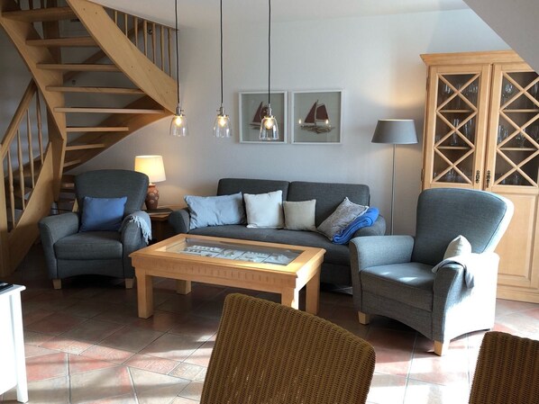 A living room with a grey sofa, two armchairs, a coffee table, and a staircase. There are framed pictures on the wall and a cabinet with glass doors.