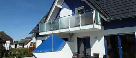 Une maison moderne à deux étages, dotée d’un balcon, d’une terrasse avec mobilier d’extérieur et surplombant un ciel d’un bleu éclatant.