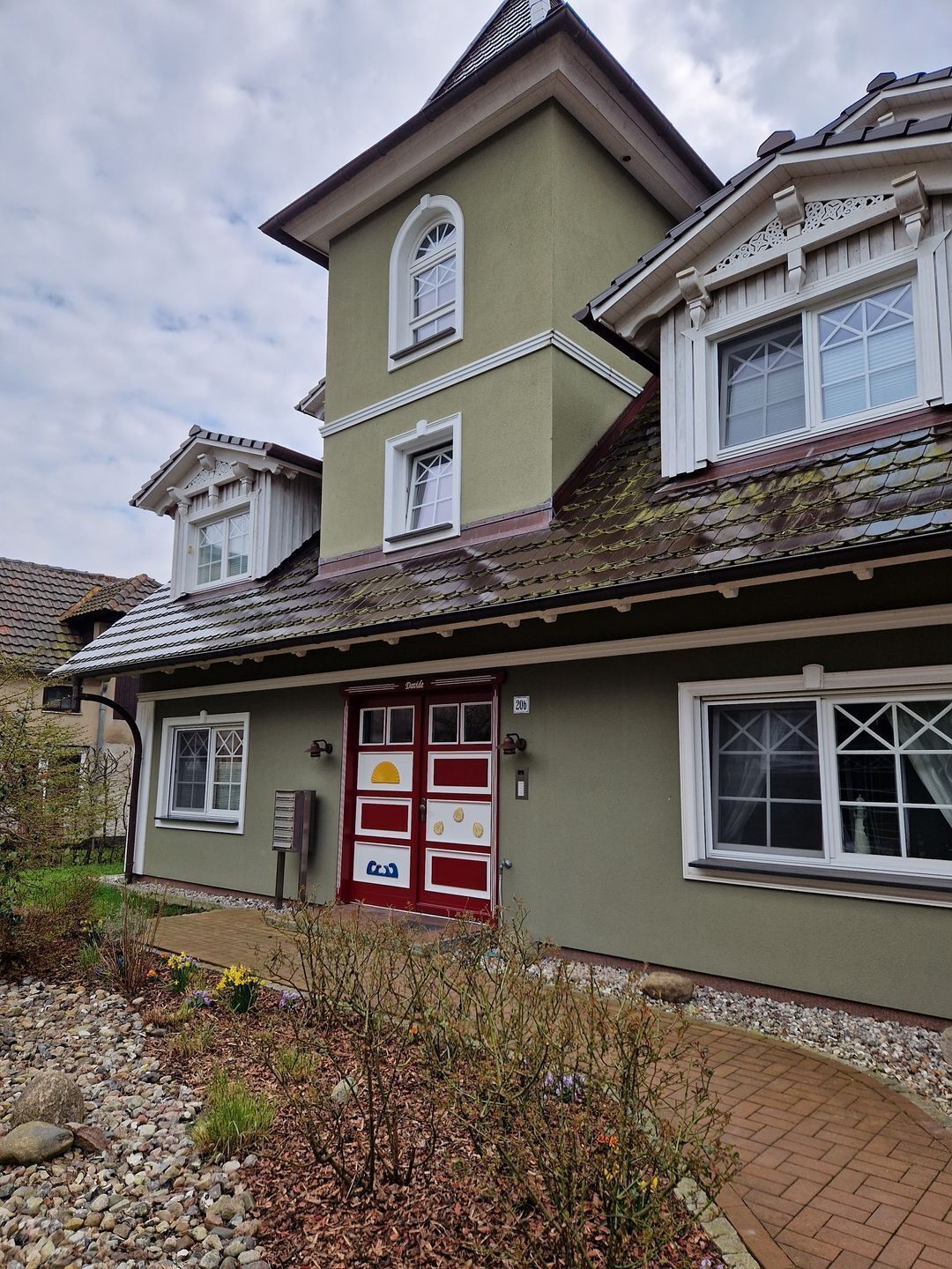 Une maison à deux étages avec une porte rouge, de nombreuses fenêtres et un petit jardin agrémenté de rochers et de plantes.