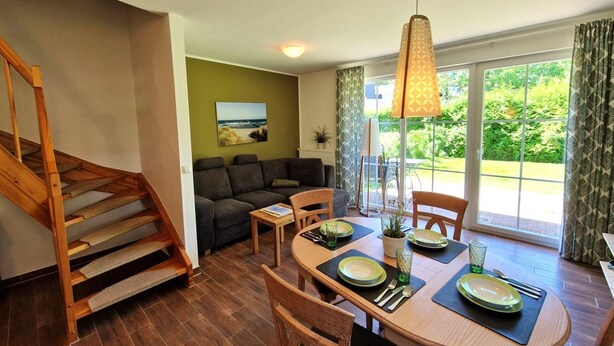 Un coin repas avec une table en bois dressée pour quatre personnes, un escalier, un canapé et une vue dégagée grâce à de grandes fenêtres.