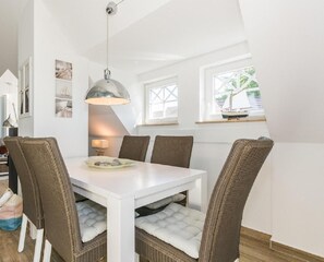 A dining area with a white table and wicker chairs, a pendant light, and a view of greenery outside.