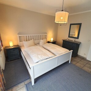 A bedroom with a white bed, two bedside tables with lamps, a dresser, a mirror, and a patterned rug.