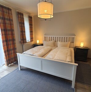 A bedroom with a white bed, two bedside tables with lamps, plaid curtains, and a textured rug.